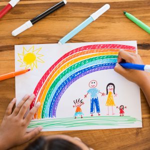 Top view of little girl’s hands painting happy family with marker on white paper. Drawing sheet with artwork by daughter on table. Little girl using marker to make family painting with rainbow.